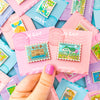 Hand holding Anne of Green Gables and Pemberley Enamel Pin set. Mix and Match Pins displayed in background are, Porthaven, Neverland, A Galaxy Far Far Away, Tulgey Woods, The Magical Castle,, Wizard of Oz, The Safari Dinosaur Island, The Wardrobe, Bag End