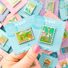 Hand holding a Mix and Match Stamp Enamel Pin Set, Porthaven, Neverland, A Galaxy Far Far Away, Tulgey Woods, The Magical Castle, Pemberley, Wizard of Oz, The Safari Dinosaur Island, The Wardrobe, Bag End, Imladris