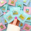 Hand holding Anne of Green Gables, Bag End, and Neverland pins on backing card. Mix and Match Pins displayed in background are Porthaven, A Galaxy Far Far Away, Tulgey Woods, The Magical Castle, Wizard of Oz, The Safari Dinosaur Island, The Wardrobe,