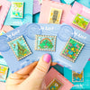 Hand holding group of Stamp Enamel Pins featuring Wizard of Oz, neverland, tulgey woods